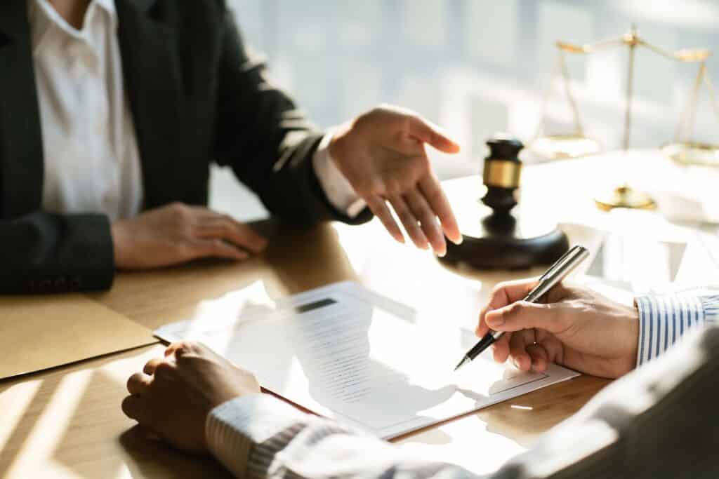 An attorney at a desk next to a gavel gesturing toward a document in front of a client holding a pen.