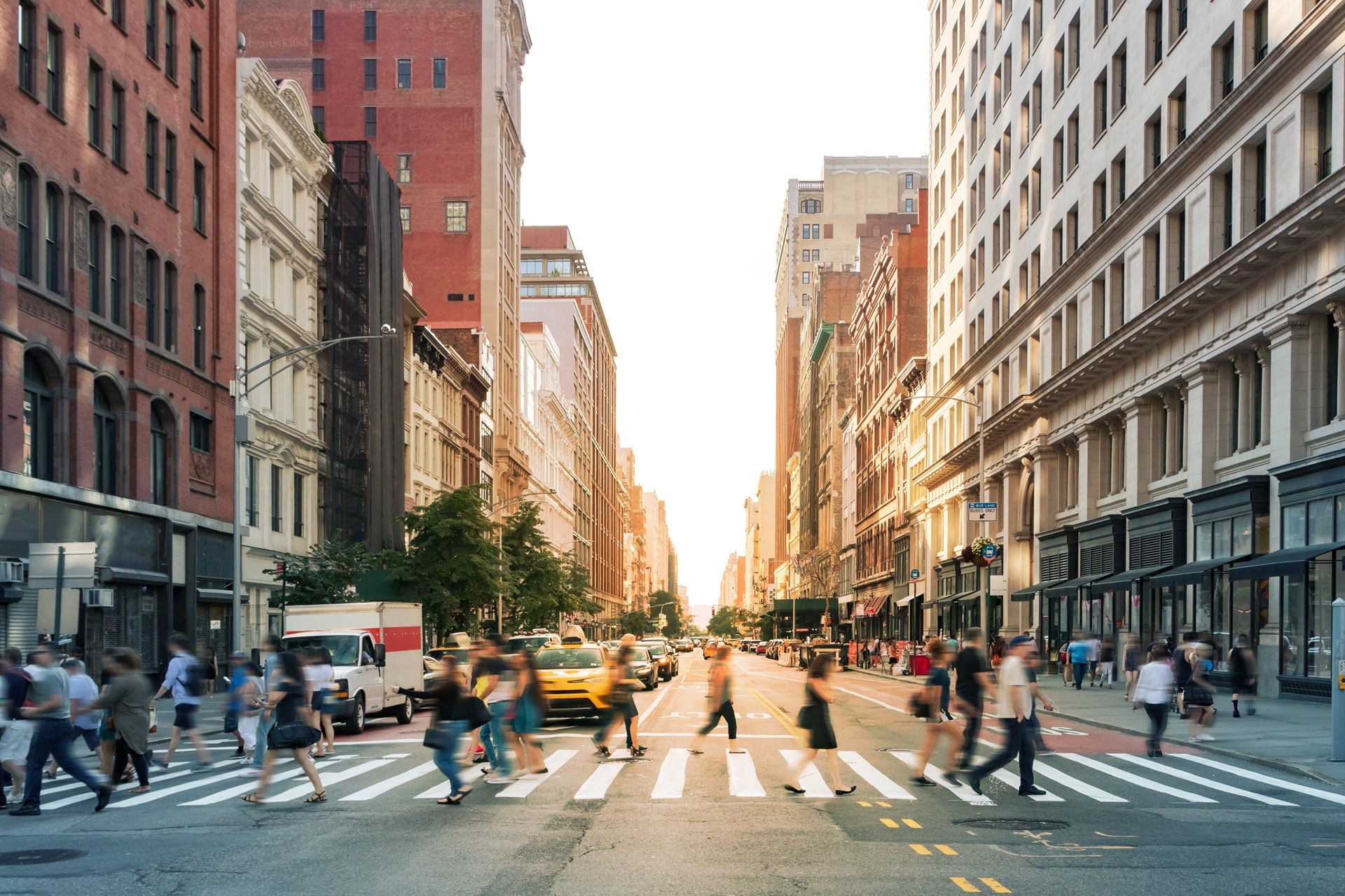 Pedestrians walking across a crosswalk in the city with the sun setting in the background.