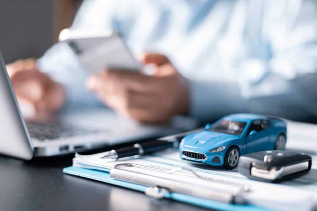 A person holding a cell phone in front of a laptop and next to a clipboard with a pen and model car on it.