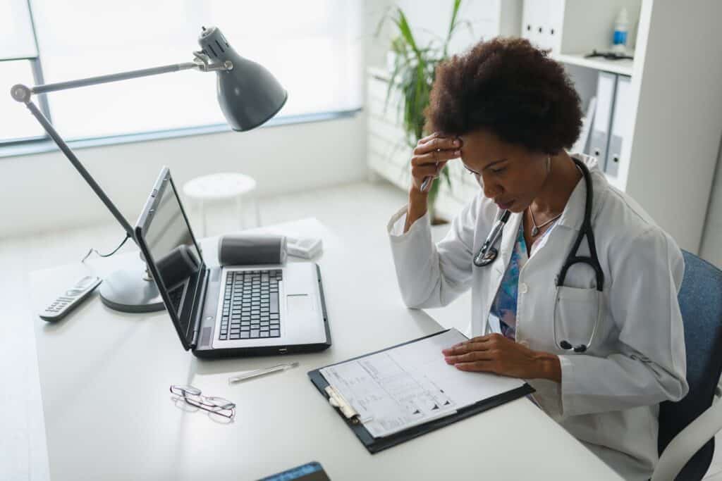 A female doctor sits behind a computer and a pad of paper looking distressed.