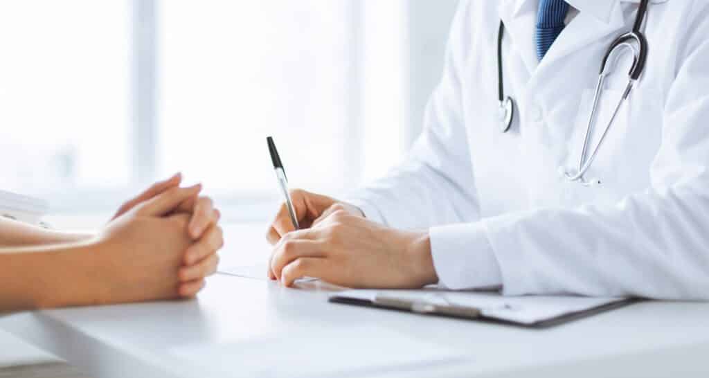 A doctor sits across from a patient at a desk holding a pen and resting their arms on paperwork.