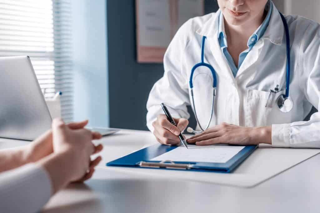A doctor writing on a clipboard while a patient sits across a desk from them with their hands held together.