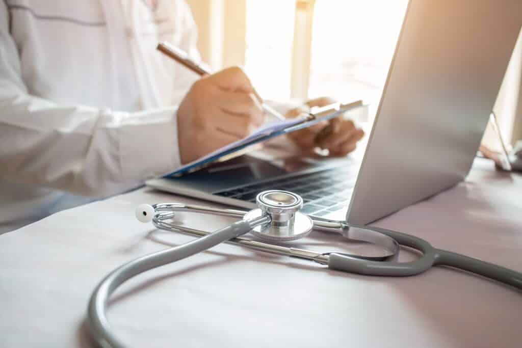 A doctor writes on a pad of paper at a desk in front of a laptop next to a stethoscope.