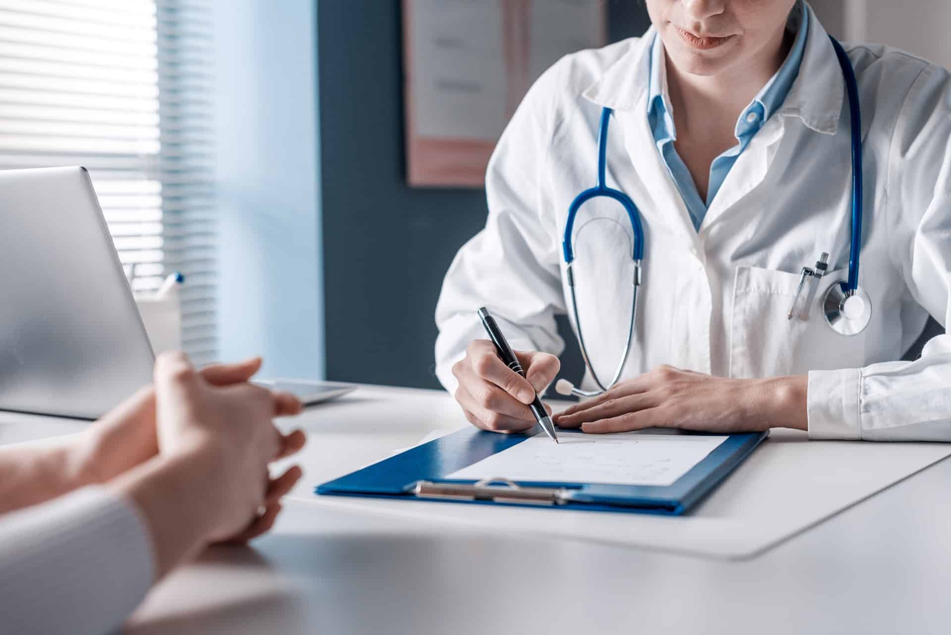 A doctor writing on a clipboard while a patient sits across a desk from them with their hands held together.