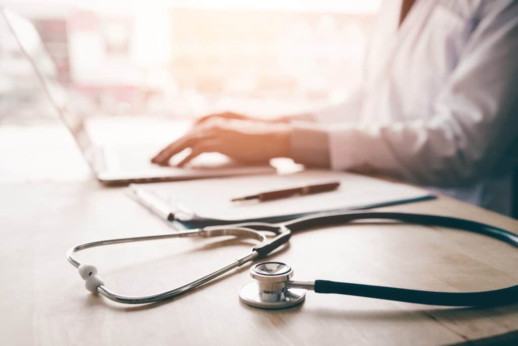 A doctor types on a laptop at a desk next to a pen resting on a pad of paper and a stethoscope.