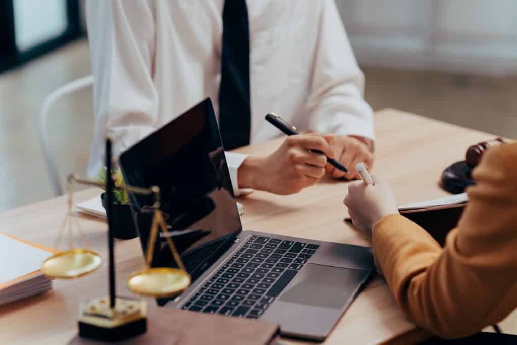 An attorney sits across a desk from a client next to a laptop and scales of justice.
