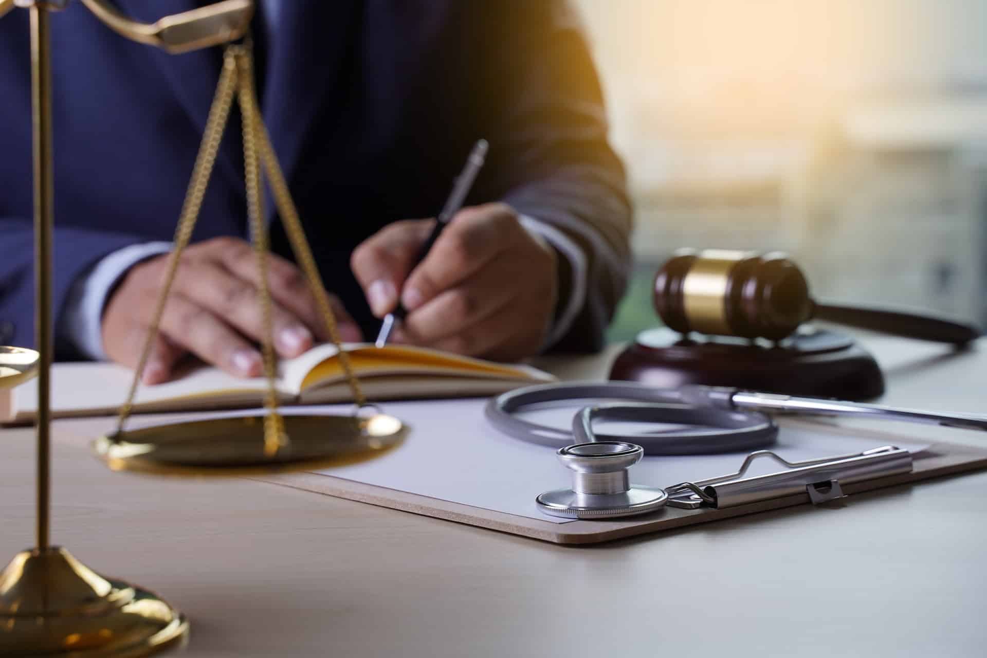 A person sitting at a desk writing on paper next to a stethoscope, gavel, and scales of justice.