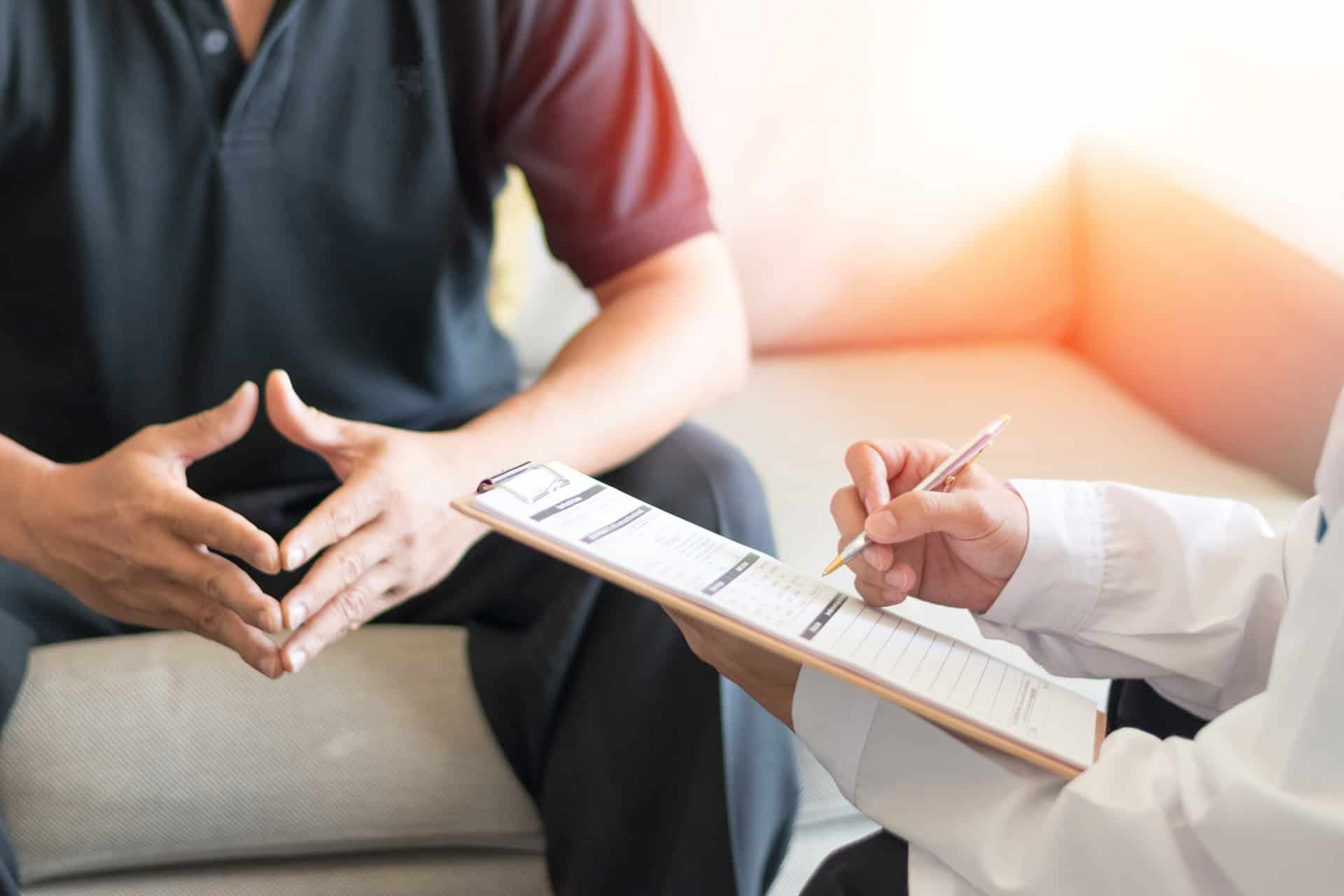 A doctor with a clipboard and pen sits in front of a patient sitting on a couch.