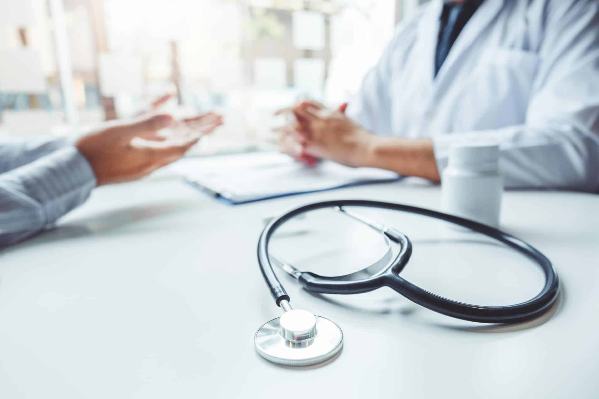 Two medical professionals sit at a white desk in front of a clipboard talking next to a stethoscope.